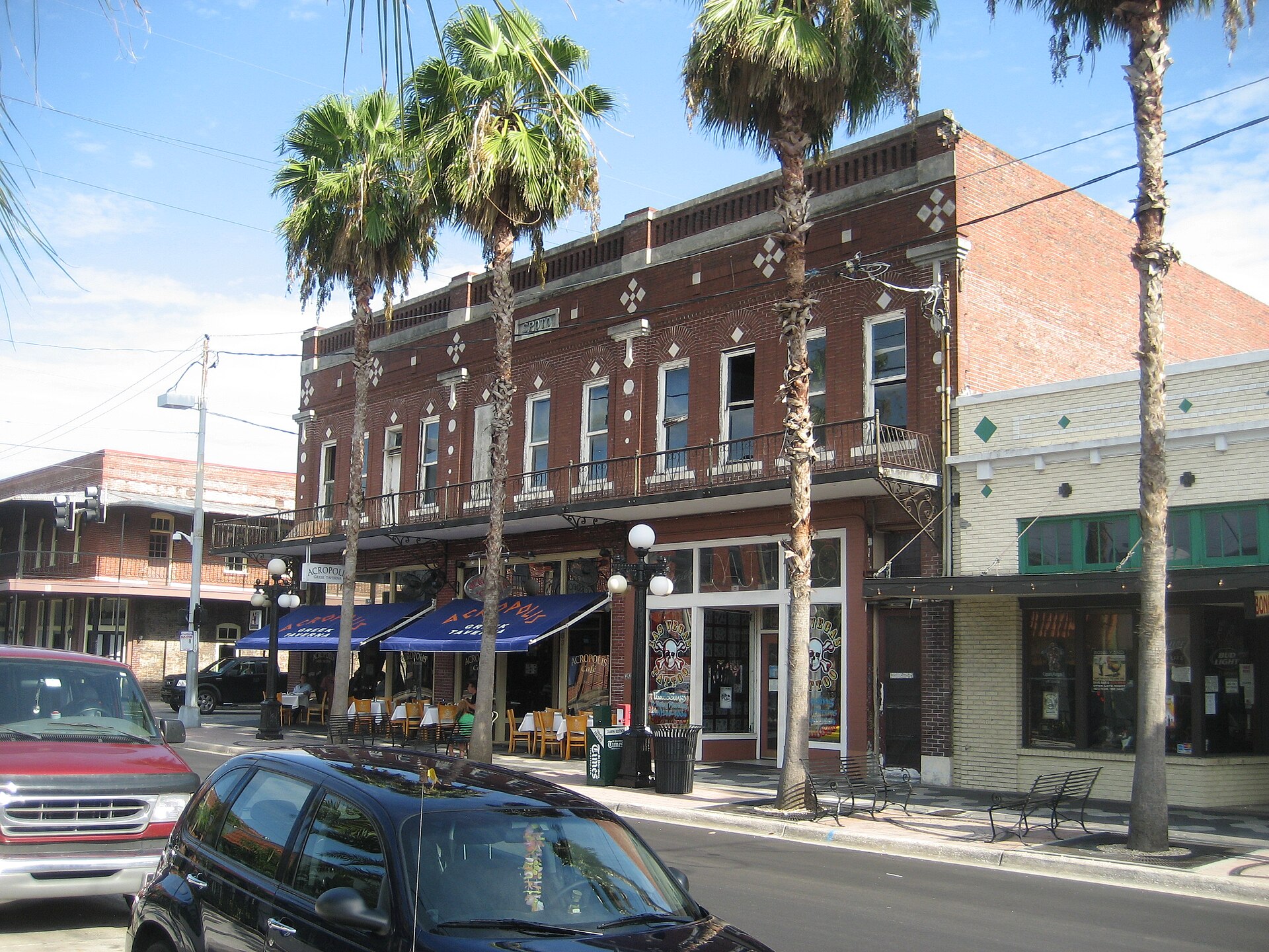 Seventh Avenue in Ybor City: two-story brick cigar-era buildings with wrought-iron balconies, palm trees and café tables under blue awnings